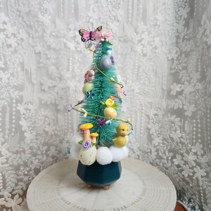 May include: A decorative Easter tree with a teal base and a green, brush-like texture. It is adorned with pastel-colored eggs, flowers, mushrooms, and a yellow duckling. A pink butterfly and bow sit atop the tree.