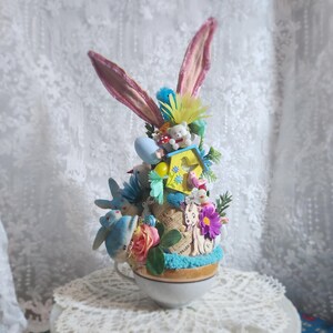 Op de afbeelding: Een grillig, op Pasen geïnspireerd decoratief theekopjesarrangement. Met konijnenoren, miniatuurvogels, een vogelhuisje en een teddybeer. De theekop is gevuld met kleurrijke bloemen, een blauwe basis en diverse decoratieve elementen. De opstelling staat tegen een kanten achtergrond.