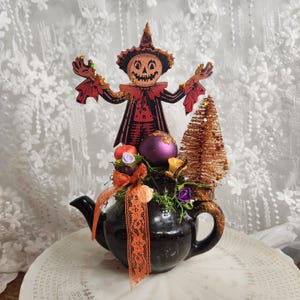 May include: A black ceramic teapot decorated with a vintage-style jack-o'-lantern cutout, a purple ornament, a small copper-coloured bottle brush tree, orange lace ribbon, and small artificial flowers, creating a festive Halloween arrangement.