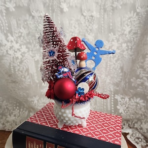 May include: A festive arrangement featuring a red bottle brush tree, red ornaments, and mushroom figurines. The arrangement is in a white hobnail vase, resting on a book with a red and white patterned cover. Decorative and colorful.