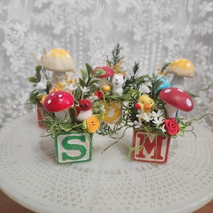 May include: A collection of miniature decorative arrangements featuring wooden blocks with letters, small figurines, and artificial flowers. The arrangements include red and yellow mushrooms, birds, and other whimsical elements, creating a charming, handcrafted aesthetic.