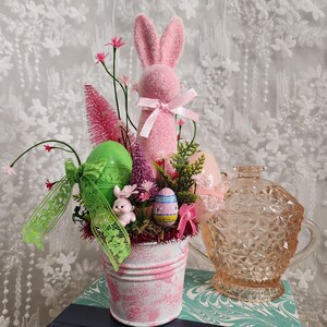 May include: An Easter-themed arrangement in a distressed pink and white bucket. It features a pink bunny, decorative eggs, and greenery. A vintage pink glass candy dish sits beside the bucket, creating a festive and decorative display.