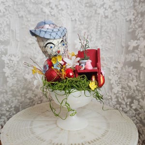 Puede incluir: Un centro de mesa caprichoso con temática de Pascua en un cuenco blanco con pedestal. Presenta una figura de muñeca con sombrero y velo azules, rodeada de huevos rojos con purpurina, flores y un banco rojo en miniatura con una figura de conejito rosa.