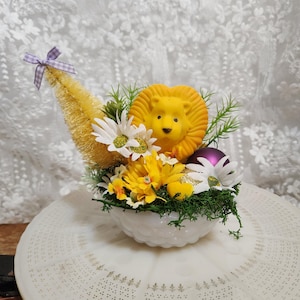 Könnte beinhalten: Dekorative Anordnung in einer weißen Schale mit einer gelben Löwenfigur, gelben Blumen, weißen Gänseblümchen, einem kleinen gelben Baum und einem lila Ornament. Die Anordnung ist mit grünem Laub und einer lila Schleife akzentuiert.
