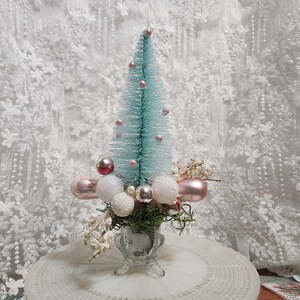 Op de afbeelding: Een kleine, turquoise kerstboom met roze en zilveren ornamenten. De boom staat in een heldere, decoratieve vaas met witte en zilveren ballen en kunstgroen. De achtergrond is een witte kanten stof.