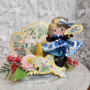 May include: A vintage paper doll in a blue and white dress, holding a fan, forms part of a decorative arrangement. The arrangement includes a cottage scene, pink flowers, and a small yellow tree. The base is silver.