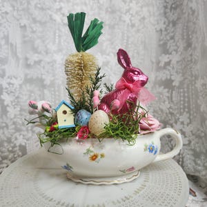Op de afbeelding: Een paasarrangement in een bloemen theekop. Het bevat een roze konijn, decoratieve eieren, een vogelhuisje en een wortelvormige decoratie. De theekop is wit met bloemmotieven, wat een lente-display creëert.