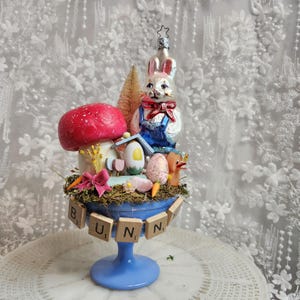 Op de afbeelding: Een grillig paasstuk met een glazen konijnenornament met een overall, een rode paddenstoel en miniatuureieren. De opstelling staat in een blauwe glazen kom met het woord "BUNNY" uitgespeld in houten tegels. Een met een ster bekroond ornament maakt de feestelijke presentatie compleet.
