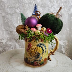 May include: A ceramic mug featuring a raised turkey relief, filled with a decorative arrangement of a dark green velvet pumpkin, a purple ornament, a small squirrel figurine, miniature mushrooms, green moss, and tiny bottle brush trees.