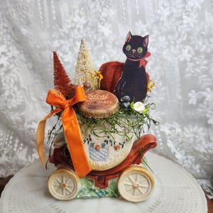 May include: A ceramic carriage-shaped planter filled with decorative elements including a black cat cutout, a round Ideal Dairy Chocolate Drink bottle cap, two small bottle brush trees, a velvet pumpkin, and an orange velvet ribbon bow.