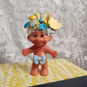 May include: A vintage troll doll with a floral headdress and a light blue bow. The doll has a smiling expression and is adorned with yellow, blue, and gold decorative elements. The doll is standing on a yellow and white patterned surface.