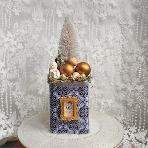 May include: A decorative Christmas arrangement in a patterned tin. The arrangement includes a small tree, gold ornaments, and a framed picture. The tin is decorated with a floral pattern in shades of gray and purple. A small figurine is also included.