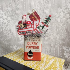 Könnte beinhalten: Eine Vintage-Currypulverdose, weihnachtlich dekoriert. Die Dose ist rot und weiß, mit dem Text "pure ground CURRY POWDER". Ein Miniatur-Weihnachtsmann in einem Schlitten, ein Geschenk und ein kleiner Weihnachtsbaum befinden sich oben.