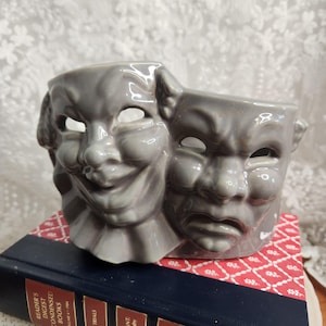 May include: A gray ceramic planter shaped like the comedy and tragedy theater masks. The masks have open eye holes and are set on a book with a red and white patterned cover. The planter is a unique decorative item.