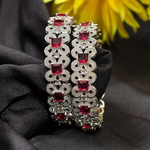 May include: Two silver bangles with a repeating openwork design, each set with square, ruby-red gemstones. The bangles rest on a black fabric, with a blurred yellow flower in the background. Jewellery.