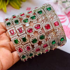 Peut inclure: Bracelets argentés ornés de pierres carrées rouges et vertes alternées. Certains bracelets présentent un motif en filigrane. Les bijoux sont présentés sur un fond floral doux, mettant en valeur les détails complexes et les couleurs vives des bracelets.