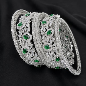 May include: A set of three silver-toned bangles featuring an intricate openwork design. Each bangle is encrusted with numerous small clear white stones and accented with oval-shaped emerald green stones arranged in a repeating floral pattern.