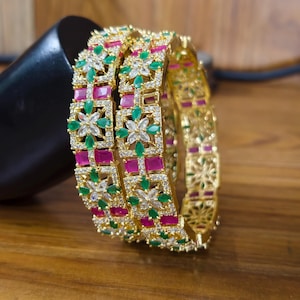 Könnte beinhalten: Zwei goldfarbene Armreifen mit einem sich wiederholenden Muster aus quadratischen Rahmen. Jeder Rahmen enthält ein zentrales Blumenmotiv aus marquise-geschliffenen grünen und klaren Steinen, abwechselnd mit rechteckigen rosa Steinen und kleinen klaren Pavé-Kristallen.
