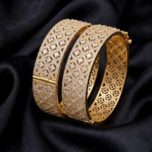 Könnte beinhalten: Zwei goldfarbene Klapparmreifen mit einer breiten, gitterartigen Oberfläche, die mit zahlreichen kleinen, klaren, quadratisch geschliffenen Steinen besetzt ist. Die Armreifen haben ein metallisches Finish und ein strukturiertes Design.