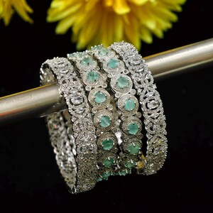 Puede incluir: Una colección de brazaletes plateados con diseños intrincados y piedras claras y de color verde azulado. Los brazaletes se exhiben en una barra plateada sobre un fondo negro, con flores amarillas en el fondo.