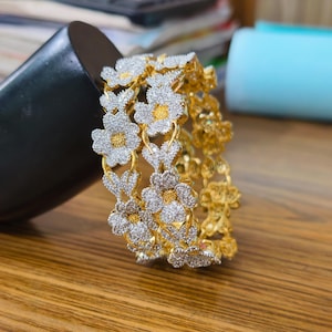 May include: A pair of gold-toned bangles featuring a repeating floral pattern. Each flower is encrusted with small, clear, sparkling stones and has a bright yellow centre. The metal structure connecting the flowers is a polished gold tone.