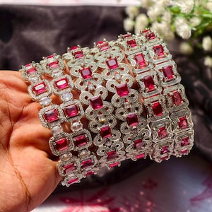Può includere: Un set di braccialetti argentati con pietre preziose quadrate rosso rubino. I braccialetti presentano un design traforato con motivi quadrati e floreali alternati. Le pietre sono incastonate in una montatura in metallo argentato. Sfondo sfocato, fiori bianchi.
