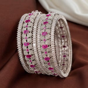 May include: A set of two silver-toned bangles featuring a central row of oval-cut pink stones interspersed with smaller round pink stones. The bangles are encrusted with numerous small clear white stones and bordered by rows of baguette-cut clear stones.