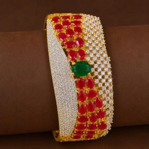 May include: A gold-toned cuff bracelet featuring a wide band encrusted with numerous small clear white stones. The design includes clusters of oval-shaped red stones and a single prominent oval green stone centered on the surface of the bracelet.