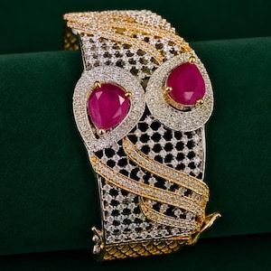 Puede incluir: Un brazalete ancho con un diseño de rejilla metálica en tonos oro y plata, incrustado con numerosas piedras pequeñas y claras. Dos piedras prominentes en forma de pera, de color rojo rosado intenso, están engastadas en el centro.