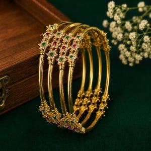Puede incluir: Un conjunto de cuatro brazaletes dorados con un motivo floral repetitivo. Cada elemento floral está incrustado con pequeñas piedras transparentes y acentuado con piedras rojas y verdes alternas. Los brazaletes tienen un acabado dorado pulido.