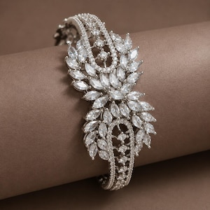Può includere: Un bracciale rigido color argento caratterizzato da una fitta disposizione di pietre trasparenti taglio marquise. Il design include due fasce curve con incastonatura a pavé che incorniciano un gruppo centrale di cristalli a forma di foglia, creando un motivo floreale simmetrico.