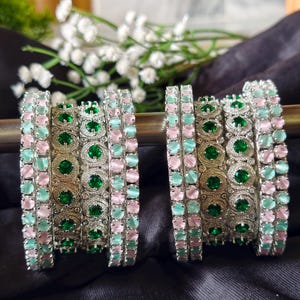 May include: Two silver bangles with alternating rows of pink, green, and blue-green gemstones. The bangles are displayed on a dark surface, with white flowers in the background. The gemstones are oval-shaped and set in a decorative silver metal.