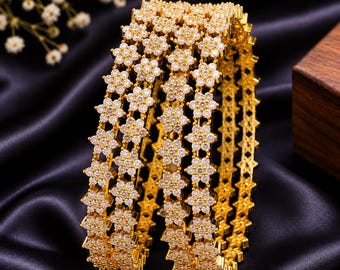 Puede incluir: Un conjunto de tres brazaletes dorados con un patrón floral repetitivo incrustado con piedras transparentes y brillantes. Cada brazalete tiene una forma circular uniforme con un acabado metálico y detalles de pequeñas flores.