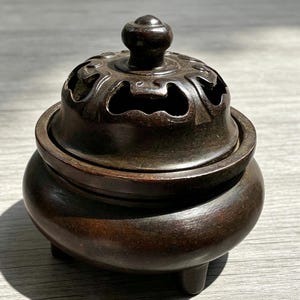 May include: A dark brown incense burner with a decorative lid featuring cut-out patterns. The burner has a rounded body and three small feet. The lid has a small knob on top. The overall design is traditional and ornate.