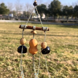 May include: A hanging mobile featuring a small wooden bar with decorative elements. The mobile includes a gourd-shaped piece, two walnut-like pieces, and two black, rounded pieces, all suspended by string. The background is a blurred outdoor scene.