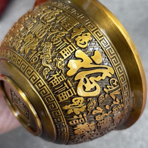 Op de afbeelding: Een bronskleurige kom met ingewikkelde reliëf Chinese karakters en bloempatronen. De buitenkant van de kom heeft gedetailleerd reliëfwerk, met een prominent centraal karakter in een contrasterende gouden tint. Het ontwerp suggereert een cultureel of decoratief item.