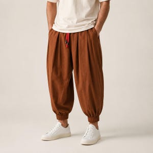 Op de afbeelding: Losvallende bruine broek met een hoge taille, diepe plooien aan de voorzijde, een elastische tailleband met een felrood trekkoord en elastische boorden bij de enkels, gecombineerd met een effen wit T-shirt en witte sneakers.