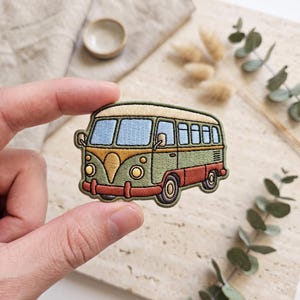 May include: An embroidered patch shaped like a vintage camper van. The van features a cream-colored roof, light blue windows, a sage green body, and a dark orange lower section with matching bumpers and wheels. The design includes detailed stitching.