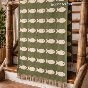 Op de afbeelding: Een groene vloerkleed met een herhalend patroon van witte vissen. Het kleed wordt getoond op een houten trap met bamboe leuningen. Het kleed heeft een gefranjerde rand en de tekst "Teppich Art Rugs" is zichtbaar.