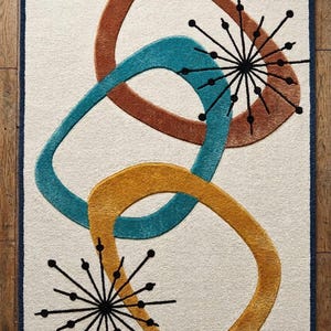 May include: A rectangular rug with a cream background and a navy blue border. The rug features overlapping abstract shapes in brown, teal, and yellow, along with black starburst designs. The text "Teppich Art Rugs" is in the top left corner.