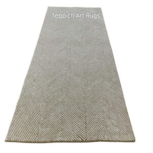 Può includere: Un tappeto rettangolare con un motivo a spina di pesce nei toni del beige e del bianco. Il tappeto ha una superficie strutturata ed è etichettato "Teppich Art Rugs" in alto al centro.