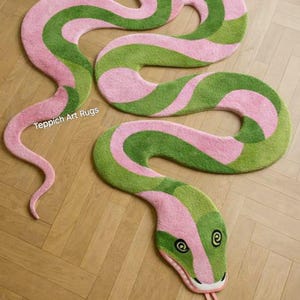 May include: Two snake-shaped rugs in pink and green. The rugs are designed to resemble a snake, with a head, body, and tail. The head has eyes and a tongue. The rugs are on a wooden floor. Text on the rug says "Teppich Art Rugs".