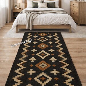 May include: A rectangular black rug with a geometric pattern in shades of tan and orange. The rug is placed in a bedroom setting, with a bed and wooden furniture in the background. The rug has a fringed edge.