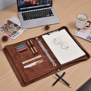 Puede incluir: Un portafolio de cuero marrón con un bloc de notas, bolígrafos y ranuras para tarjetas. Una computadora portátil, una taza de café y un periódico están sobre el escritorio de madera. Gafas redondas descansan sobre el bloc de notas.