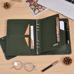 May include: Open green leather binder with multiple pockets, a zipper compartment, and silver ring binder. A black pen, glasses, and a smartphone are on the wooden surface. A laptop and a magazine are in the background.