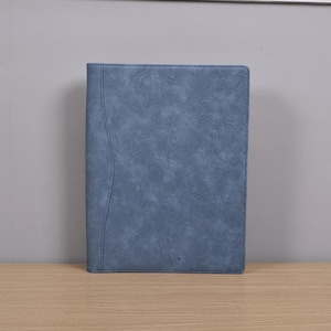 Op de afbeelding: Een blauwe portfolio met leerlook en een getextureerde afwerking. De omslag heeft een gestikte rand en een gebogen designelement aan de linkerkant. De rechthoekige portfolio is gesloten, wat suggereert dat deze documenten of een notitieblok kan bevatten.