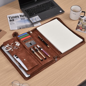 May include: A brown leather portfolio case with multiple compartments, including card slots, pen holders, and a notepad. The case is open, revealing a laptop, pens, and a smartphone. A laptop, newspaper, and mug are also visible.