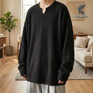 Op de afbeelding: Zwart shirt met lange mouwen, geribbelde textuur en een V-hals. Het shirt heeft een relaxte pasvorm en lange mouwen. Het KRIMSANOVA-logo is zichtbaar in de rechterbovenhoek. Het shirt wordt gecombineerd met een grijze joggingbroek.