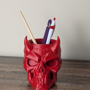 May include: A red skull-shaped pen holder with horns, holding a wooden pencil, a purple pen, and a red and white pen. The pen holder is made of a glossy material and sits on a wooden surface.