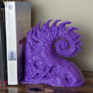 May include: A purple, 3D-printed bookend with a detailed, organic design. The bookend has a swirling, horn-like shape and is placed next to two books. The bookend is made of plastic and is designed to hold books upright.